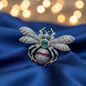 Puede incluir: Broche de abeja con incrustaciones de diamantes, con detalles de esmeraldas y rubíes. El broche presenta un diseño detallado con alas brillantes y una piedra central de esmeralda. El cuerpo de la abeja está adornado con rubíes, las patas son doradas.