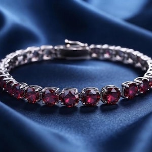 May include: A tennis bracelet featuring a continuous row of round-cut, deep red gemstones set in a polished silver-toned metal frame. The bracelet includes a secure box clasp mechanism and displays a consistent, symmetrical design throughout.