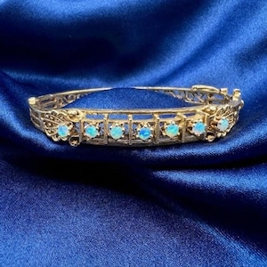 May include: A gold-toned bracelet with a filigree design and six oval, light blue gemstones. The bracelet is set against a deep blue, silky fabric, highlighting its intricate details and vintage style.
