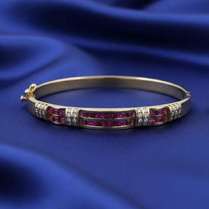 May include: A gold bangle bracelet featuring two rows of square-cut red gemstones and small white gemstones. The bracelet is set against a blue background.