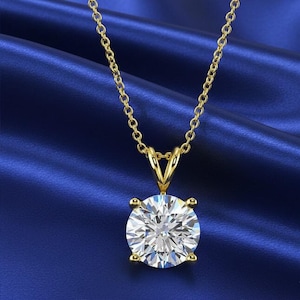 May include: A gold-colored necklace with a large, round, clear gemstone pendant. The pendant is set in a four-prong setting and hangs from a delicate chain. The background is a deep blue fabric.