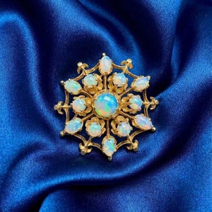 Op de afbeelding: Een goudkleurige broche met een sterrenpatroon, met een centrale opaal en kleinere opalen. De opalen vertonen iriserende kleuren. De broche ligt op een diepblauwe satijnen stof.