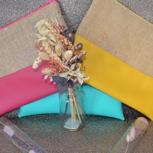 May include: Two burlap and leather zipper pouches, one pink and one yellow, are displayed with a bouquet of dried flowers in a glass jar. A turquoise cushion is beneath the pouches. Two glass tubes with dried flowers are in the foreground.