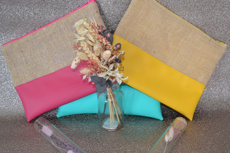 May include: Two burlap and leather zipper pouches, one pink and one yellow, are displayed with a bouquet of dried flowers in a glass jar. A turquoise cushion is beneath the pouches. Two glass tubes with dried flowers are in the foreground.