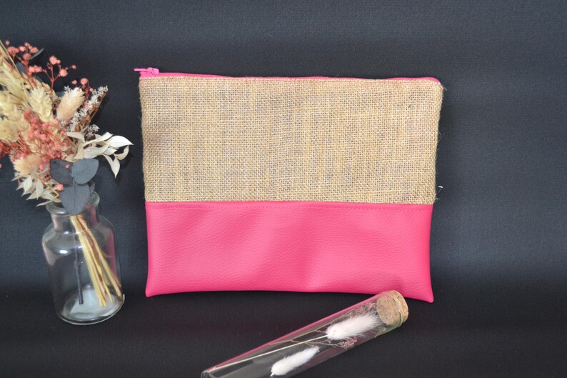 May include: A rectangular pouch with a pink base and a tan burlap top, secured with a pink zip. A clear glass vial with white decorative elements and a small glass vase with dried flowers are in the foreground.