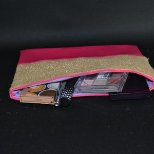 May include: A pink and gold cosmetic bag with a zip, filled with makeup products. The bag has a pink body with a gold sequined band across the top. Inside are eyeshadow palettes, a mascara tube, and other beauty items.
