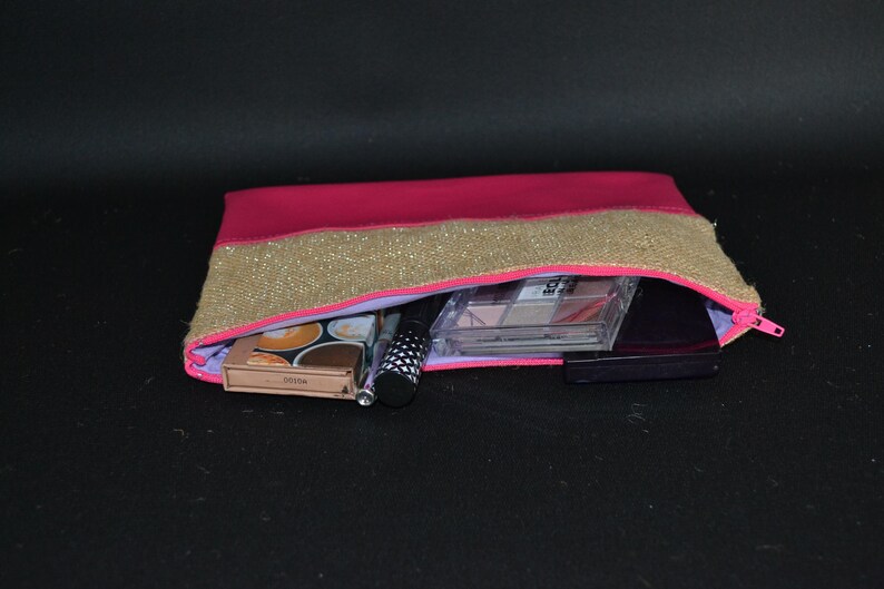 May include: A pink and gold cosmetic bag with a zip, filled with makeup products. The bag has a pink body with a gold sequined band across the top. Inside are eyeshadow palettes, a mascara tube, and other beauty items.