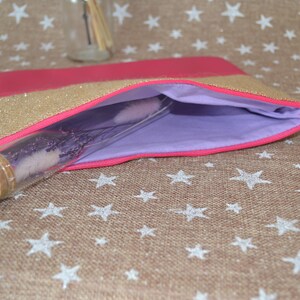 May include: A pink and gold glitter clutch bag with a lavender interior. A clear glass bottle with a cork stopper is partially inside the bag. The bag is on a tan surface with white star patterns.