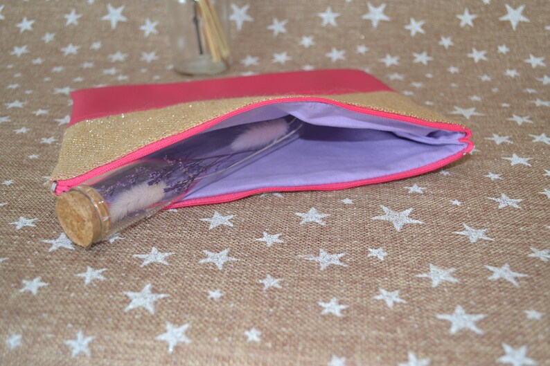 May include: A pink and gold glitter clutch bag with a lavender interior. A clear glass bottle with a cork stopper is partially inside the bag. The bag is on a tan surface with white star patterns.