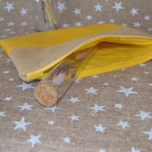 May include: A yellow and hessian pouch with a gold zip, open to show a glass vial with a cork stopper. The vial contains dried flowers and a pink stone. The pouch is on a brown surface with white star patterns.