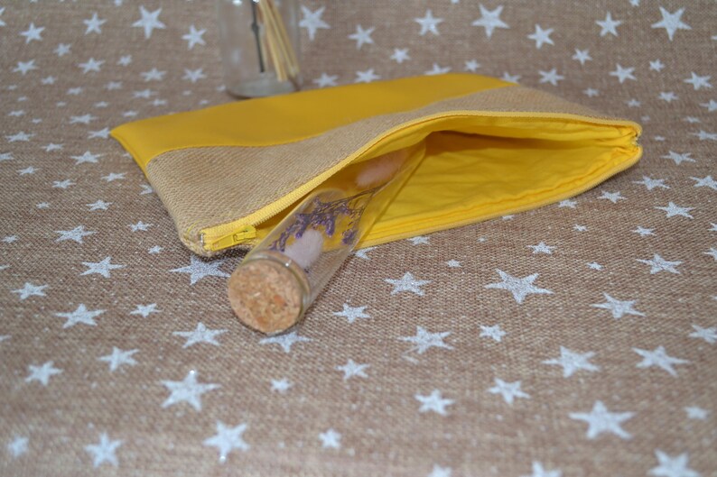 May include: A yellow and hessian pouch with a gold zip, open to show a glass vial with a cork stopper. The vial contains dried flowers and a pink stone. The pouch is on a brown surface with white star patterns.