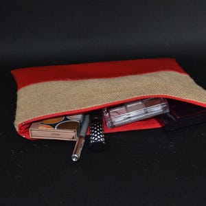 May include: A red and beige burlap cosmetic bag with a red zip, partially open to show makeup items. The bag contains eyeshadow palettes, a pen, mascara, and other beauty products. The background is black.