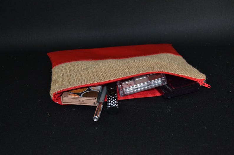 May include: A red and beige burlap cosmetic bag with a red zip, partially open to show makeup items. The bag contains eyeshadow palettes, a pen, mascara, and other beauty products. The background is black.