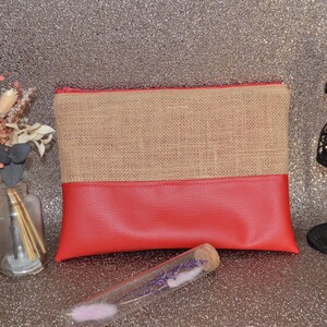 May include: A rectangular clutch bag with a burlap top and a red bottom. A black decorative mannequin and a small glass bottle with dried flowers are on the sides. A glass tube with lavender is in front.