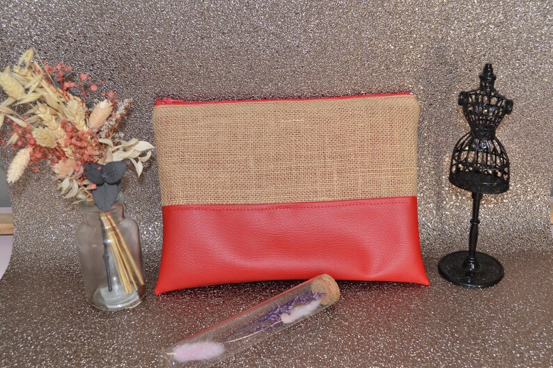 May include: A rectangular clutch bag with a burlap top and a red bottom. A black decorative mannequin and a small glass bottle with dried flowers are on the sides. A glass tube with lavender is in front.