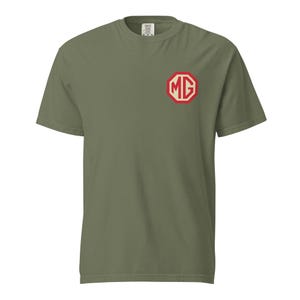 Puede incluir: Camiseta verde oliva con el logotipo MG rojo y blanco en el pecho izquierdo. El logotipo tiene forma octogonal con las letras "MG" en su interior. La camiseta está hecha de un material suave y cómodo.