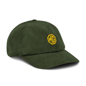 May include: A dark green corduroy baseball cap with a curved brim. The cap features a yellow and gold embroidered MG logo on the front, and a small button on top. The hat is a classic style.