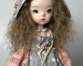 Può includere: Una bambola con un'espressione serena, che indossa un vestito grigio a pois e un cappello abbinato. Il cappello è adornato con accenti rosa e un dettaglio floreale. La bambola ha lunghi capelli castani ondulati e una piccola borsa intrecciata.