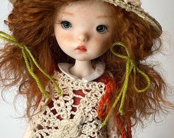 Può includere: Una bambola con capelli rossi e occhi blu indossa un cappello e un vestito all'uncinetto. Il cappello è decorato con accenti colorati. L'abito è beige con un motivo floreale. Accenti di filo verde e arancione accentuano l'outfit. La bambola ha un incarnato chiaro e guarda avanti.