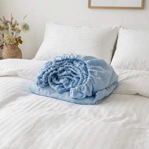 以下が含まれることがあります： 白いストライプのベッドカバーの上に、折りたたまれた水色のフィットシーツが置かれています。シーツの後ろには、白いストライプの枕が2つあります。「LINEN」と書かれた額入りのサインが背景に見えます。