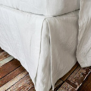 May include: A white bed skirt made of woven fabric with a visible texture. The bed skirt features a tailored design with a pleated corner detail that hangs down from the edge of the mattress, covering the lower portion of the bed frame structure.