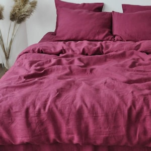May include: A bed dressed with a deep magenta linen duvet cover and pillowcases. The bedding has a slightly rumpled appearance, suggesting a soft, comfortable texture. The overall aesthetic is simple and elegant, suitable for a bedroom.