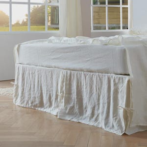 May include: A bed with white linen bedding, including a fitted sheet and a ruffled bed skirt. The bed skirt has tie closures. The room has large windows with sheer curtains, and the floor is light-colored wood.