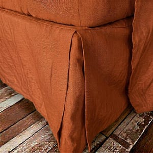 May include: A rust-colored linen bed skirt featuring a tailored box pleat design. The fabric has a visible woven texture and a matte finish, draping downward from the base of a piece of furniture to create a clean, structured appearance.