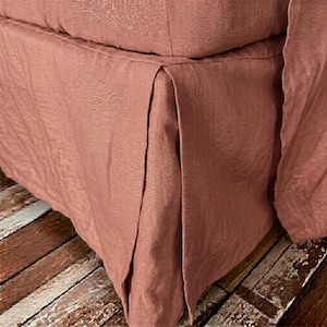 May include: A dusty rose colored bed skirt made of a textured fabric. The bed skirt features a tailored design with a clean, straight drop and a single box pleat detail visible at the corner, showcasing the woven texture of the material.
