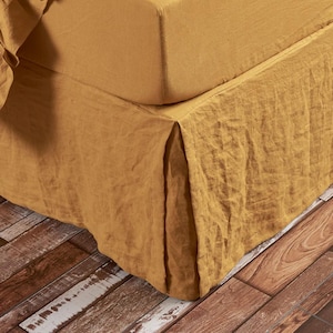 May include: Mustard-colored linen bed skirt with a pleated design. The bed skirt is draped over a bed frame, with a fitted sheet visible. The fabric has a textured appearance, and the overall aesthetic is rustic and cozy.