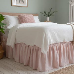 May include: A bed with a white comforter and a pink bed skirt. A fluffy pink pillow rests on the bed, and a beige throw blanket is draped over the foot of the bed. A potted plant in a woven basket sits nearby.