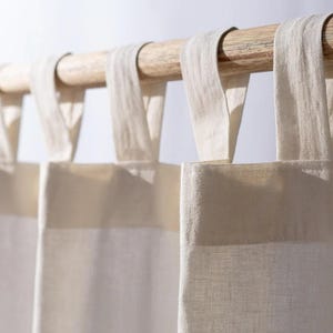 May include: Close-up of cream-coloured linen curtains suspended from a natural wood rod. The curtains feature a simple, minimalist design with fabric loops for hanging. The neutral colour palette and natural materials create a calming, airy aesthetic, suitable for home decor.