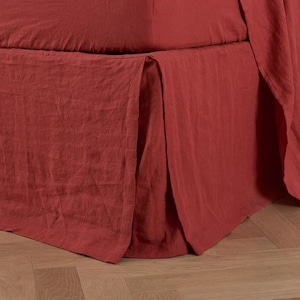 May include: A close-up of a bed with a rust-colored bed skirt and fitted sheet. The fabric has a textured appearance and drapes over the bed frame. The bed skirt has a pleated design, adding a touch of elegance to the bedding.
