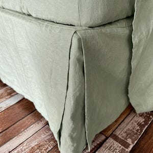 May include: A sage green bed skirt made of textured linen fabric. The bed skirt features a tailored design with a crisp box pleat at the corner, providing a clean and structured appearance that drapes smoothly along the side of the bed frame.
