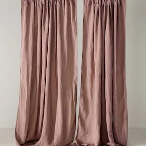 Puede incluir: Dos cortinas de lino color rosa empolvado colgadas de una barra de madera. Las cortinas están recogidas en la parte superior, creando pliegues suaves que caen hacia abajo. La tela parece ligeramente texturizada, lo que se suma a la estética natural de las cortinas. El fondo es una pared blanca lisa.