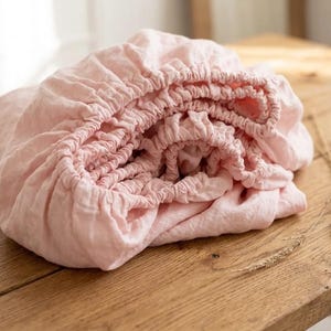 May include: A light pink fitted sheet, neatly folded on a wooden surface. The sheet has an elasticized edge and a soft, textured appearance. The bedding is made of a lightweight fabric, perfect for a bedroom.