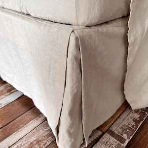 May include: Close-up of a beige linen bed skirt with a tailored pleated design. The fabric has a natural, textured appearance. The bed skirt is draped over a bed frame, with a wooden floor visible in the background.