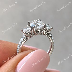 May include: A silver-toned three-stone ring featuring a large round-cut centre stone flanked by two smaller round-cut stones, all set in a prong setting. The band is adorned with a row of small pavé-set stones along the visible side of the shank.