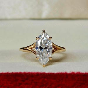 May include: A gold ring featuring a single marquise-cut clear gemstone held by six prongs. The band has a split shank design that tapers toward the center stone, creating a sleek and polished appearance for this piece of fine jewelry.