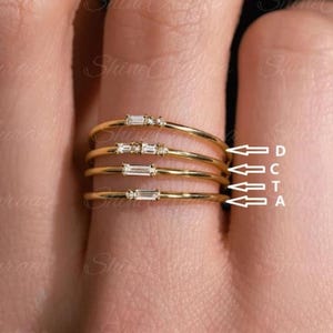 May include: Four delicate gold rings are stacked on a finger. Each ring features small, sparkling gemstones. The rings are labelled A, C, T, and D. The brand name "Shine Auraa" is visible in the background.