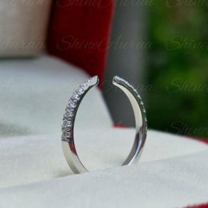 May include: A silver-toned open ring featuring a row of small, clear, round-cut stones set along one side of the band. The ring is positioned upright in a red and white jewelry box with the text ShineAuraa visible in the background.