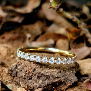 May include: A gold band ring featuring a continuous row of small, clear, round-cut stones set along the top half of the metal band. The ring is positioned horizontally, showcasing the uniform arrangement of the sparkling stones against the metal.