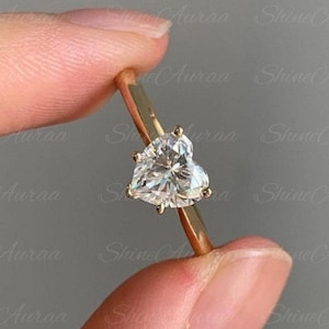 May include: A gold ring featuring a heart-shaped diamond. The diamond is clear and faceted, catching the light. The ring's band is a thin, polished gold. The ring is held between two fingers against a neutral background.