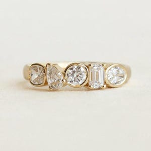 May include: A gold ring featuring five clear gemstones of varying shapes: cushion, pear, round, emerald, and round. The gemstones are set in a gold band, creating a delicate and elegant design. The ring is displayed on a white background.