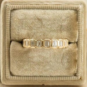 May include: A gold band ring featuring a continuous row of seven rectangular emerald-cut clear stones set in a bezel setting. The ring is positioned horizontally, showcasing the uniform metal frame and the clear, faceted stones aligned side by side.