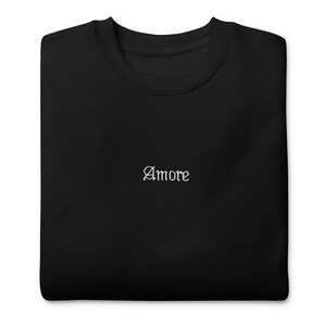 Könnte beinhalten: Schwarzes Sweatshirt mit Rundhalsausschnitt und dem Wort "Amore" in weißer Schrift auf der Brust gestickt. Das Sweatshirt ist aus einem weichen Material gefertigt und hat eine klassische, entspannte Passform. Ein schlichtes, aber stilvolles Design.