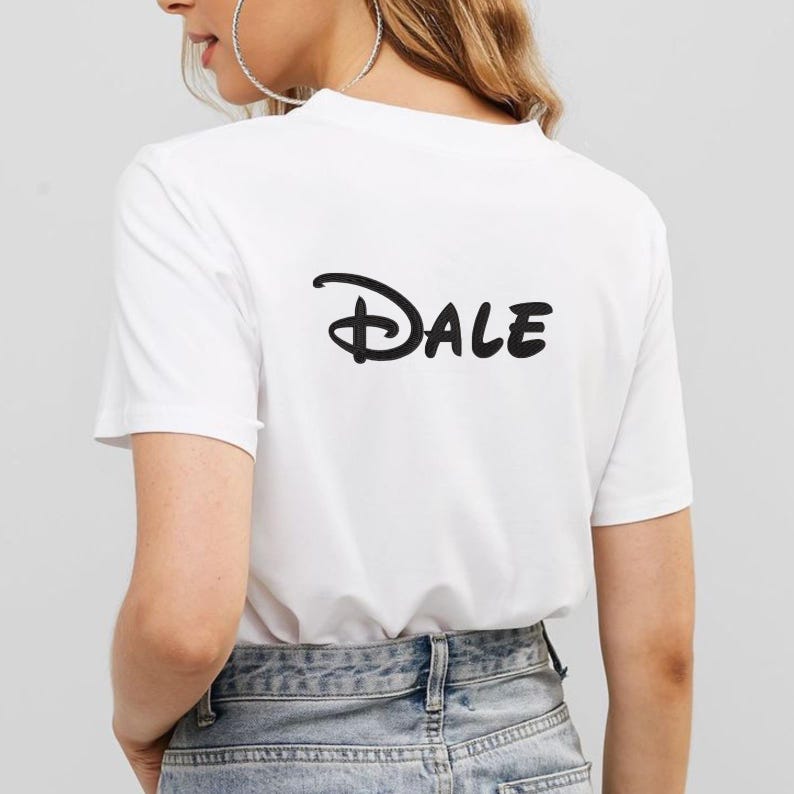 May include: White t-shirt with the name "Dale" printed in black, stylized script on the back. The shirt is a short-sleeved crew neck style. The person is wearing blue jeans and large silver hoop earrings.