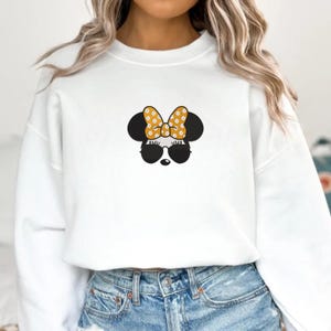 May include: White sweatshirt featuring a Minnie Mouse design. The design includes black mouse ears, sunglasses, and a yellow bow with white polka dots. The sweatshirt is paired with blue denim jeans.