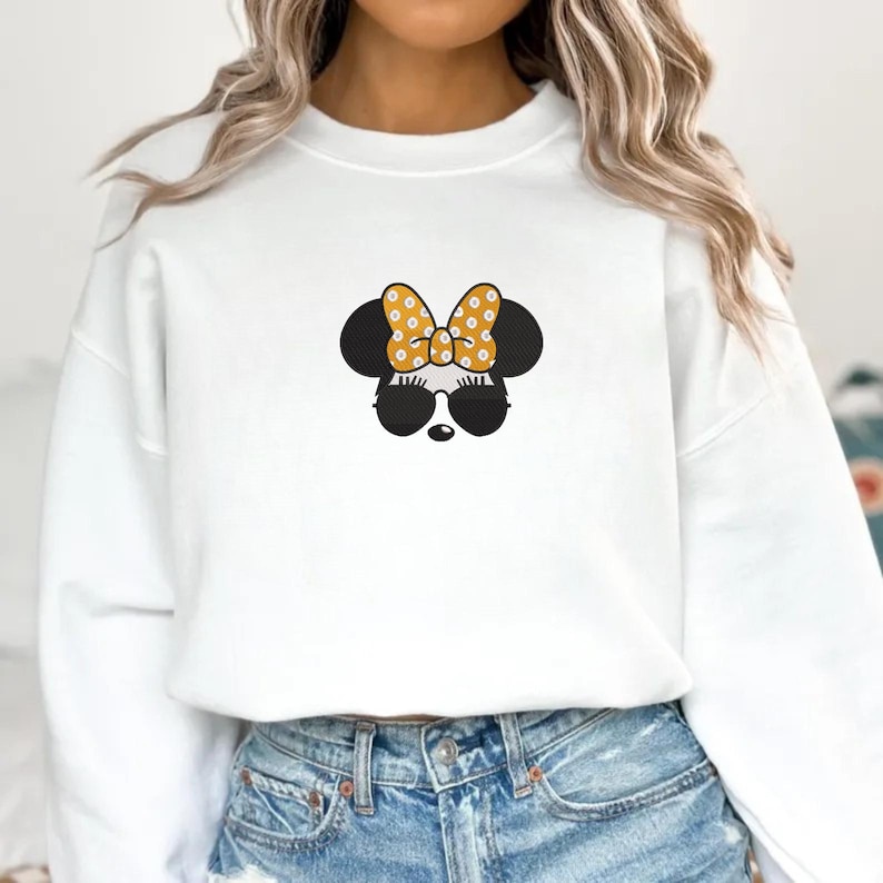 May include: White sweatshirt featuring a Minnie Mouse design. The design includes black mouse ears, sunglasses, and a yellow bow with white polka dots. The sweatshirt is paired with blue denim jeans.