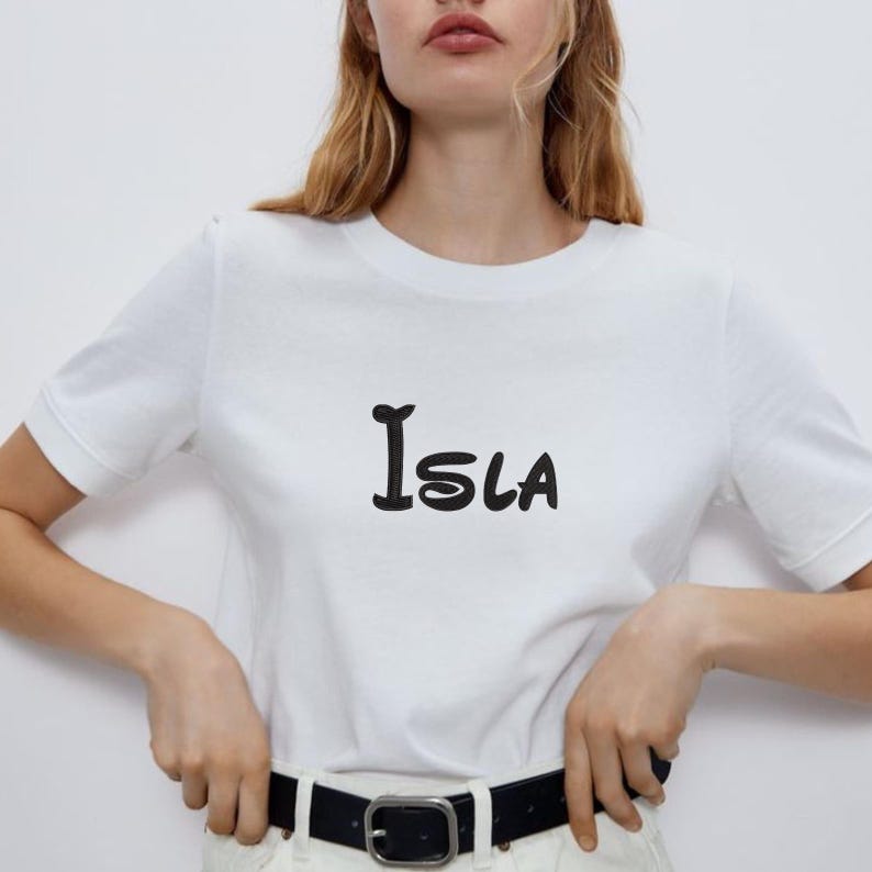 May include: White t-shirt with the name "Isla" printed in black, featuring a stylized font. The shirt has short sleeves and a crew neck. The model is wearing a black belt and white pants.